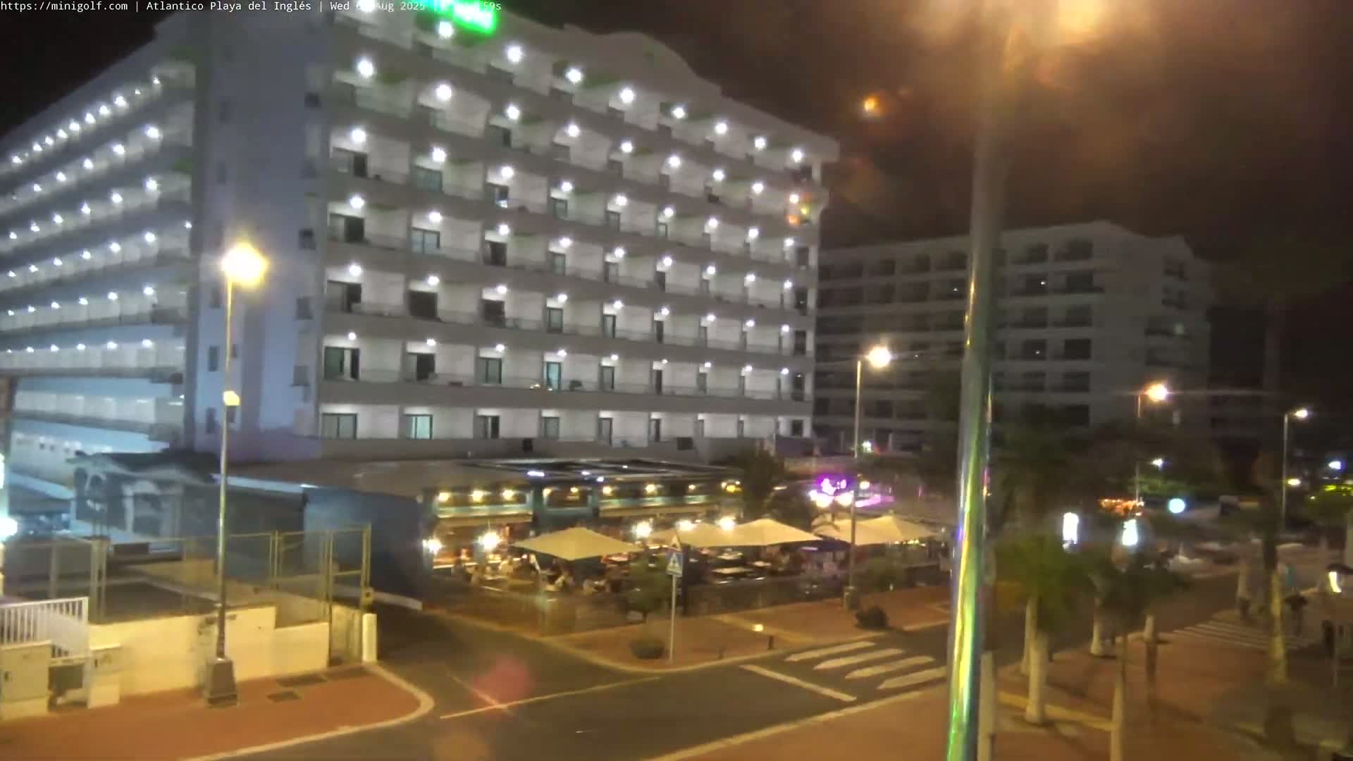 A brightly lit, multi-story hotel stands at night next to an outdoor restaurant with patrons seated under umbrellas, all under a dark sky.