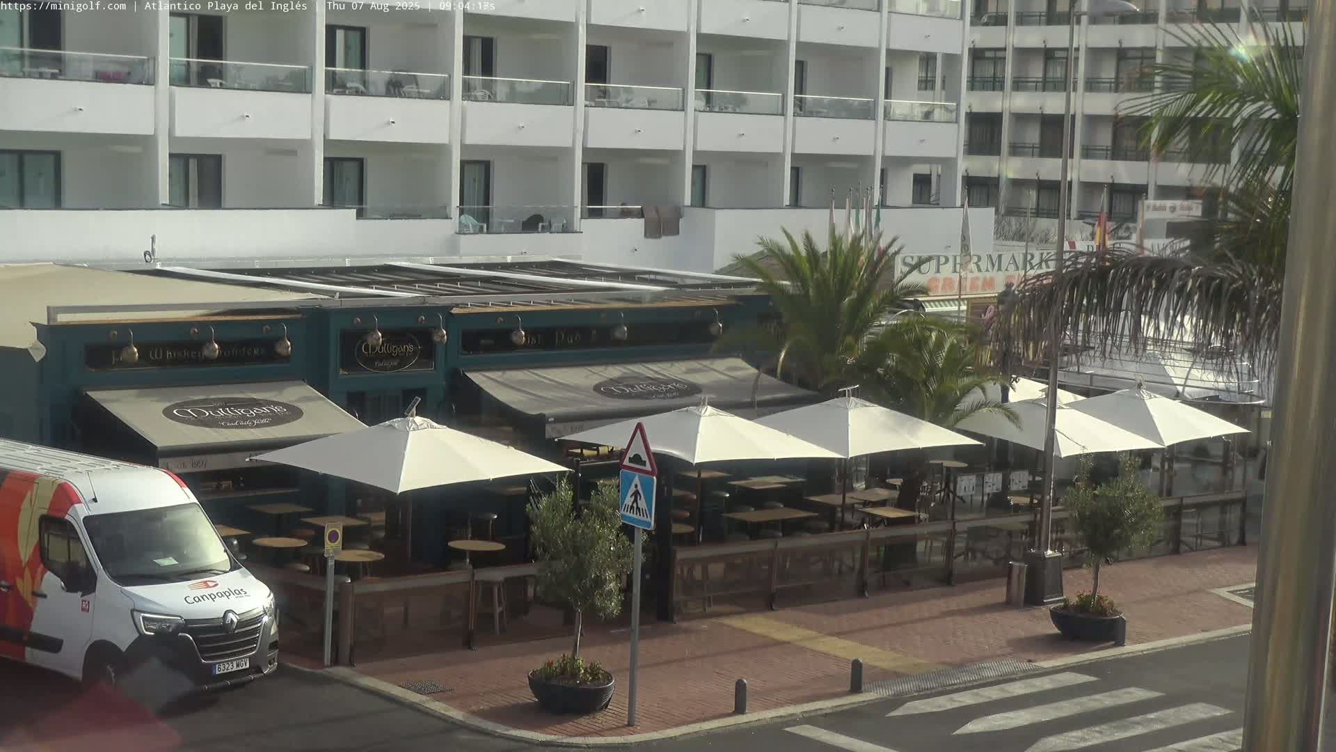 A street-level view shows a row of outdoor restaurant seating under white umbrellas, next to a building with a sign reading "Mulligans," and a van parked nearby on a sunny day.