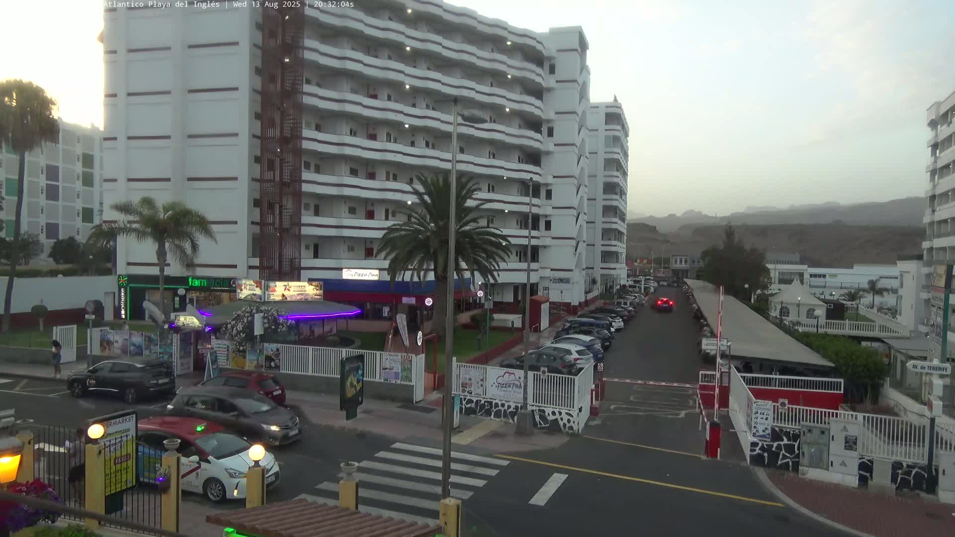Under a dusky sky, a multi-story building complex is partially visible, with cars parked in a lot and several vehicles driving on a street in front of it.