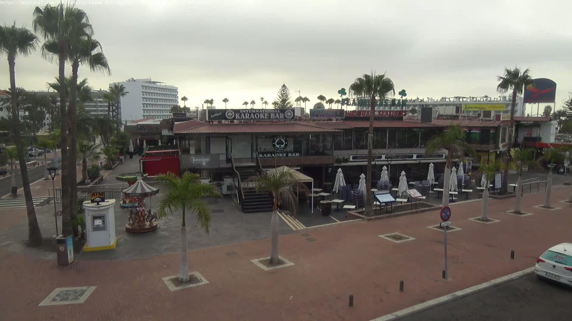 A plaza with palm trees, a carousel, several restaurants and bars, and a hotel in the background under an overcast sky.