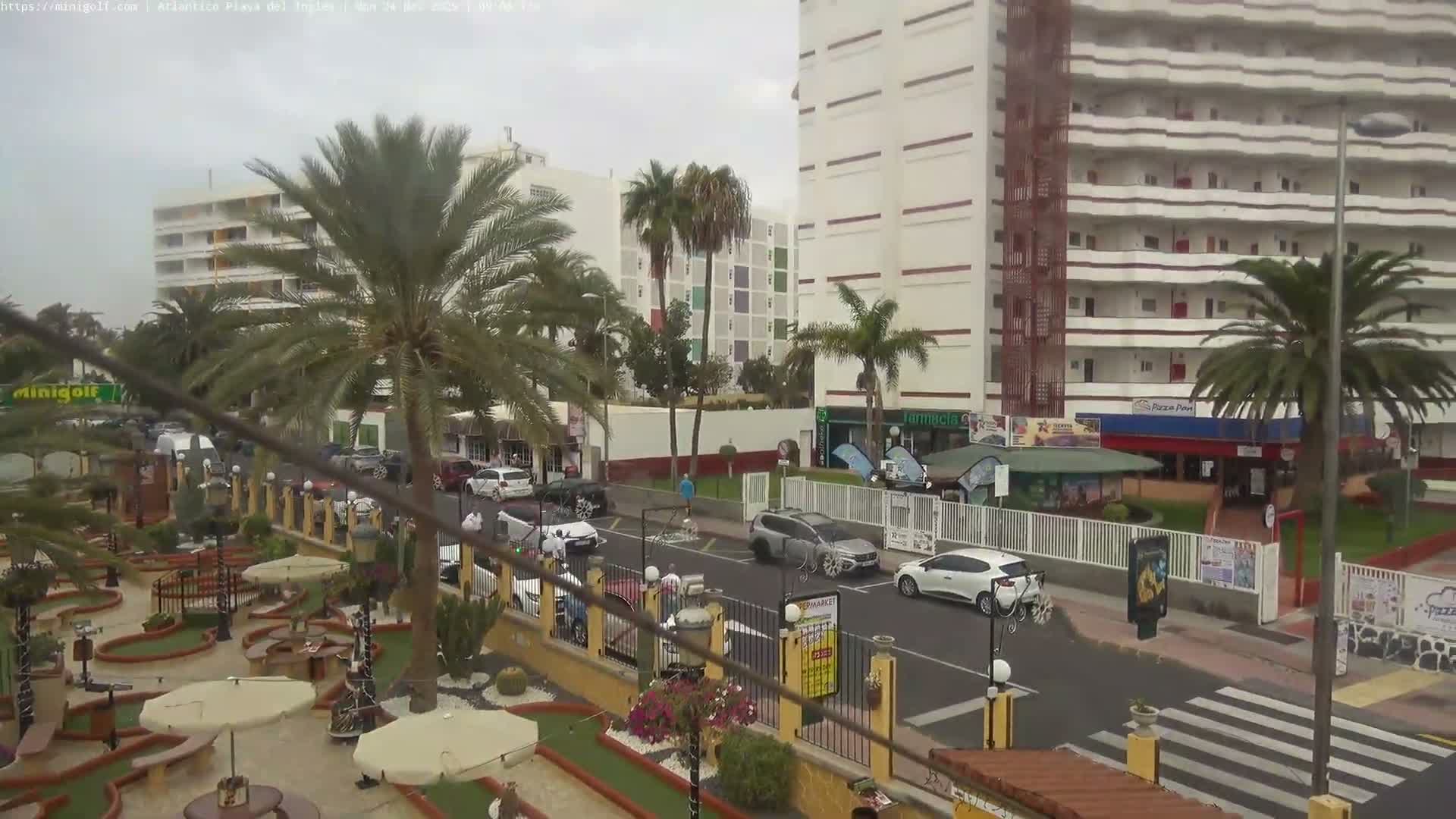 An outdoor mini-golf course with palm trees and white umbrellas occupies the foreground, facing a street with cars and pedestrians, backed by multi-story buildings under an overcast sky.