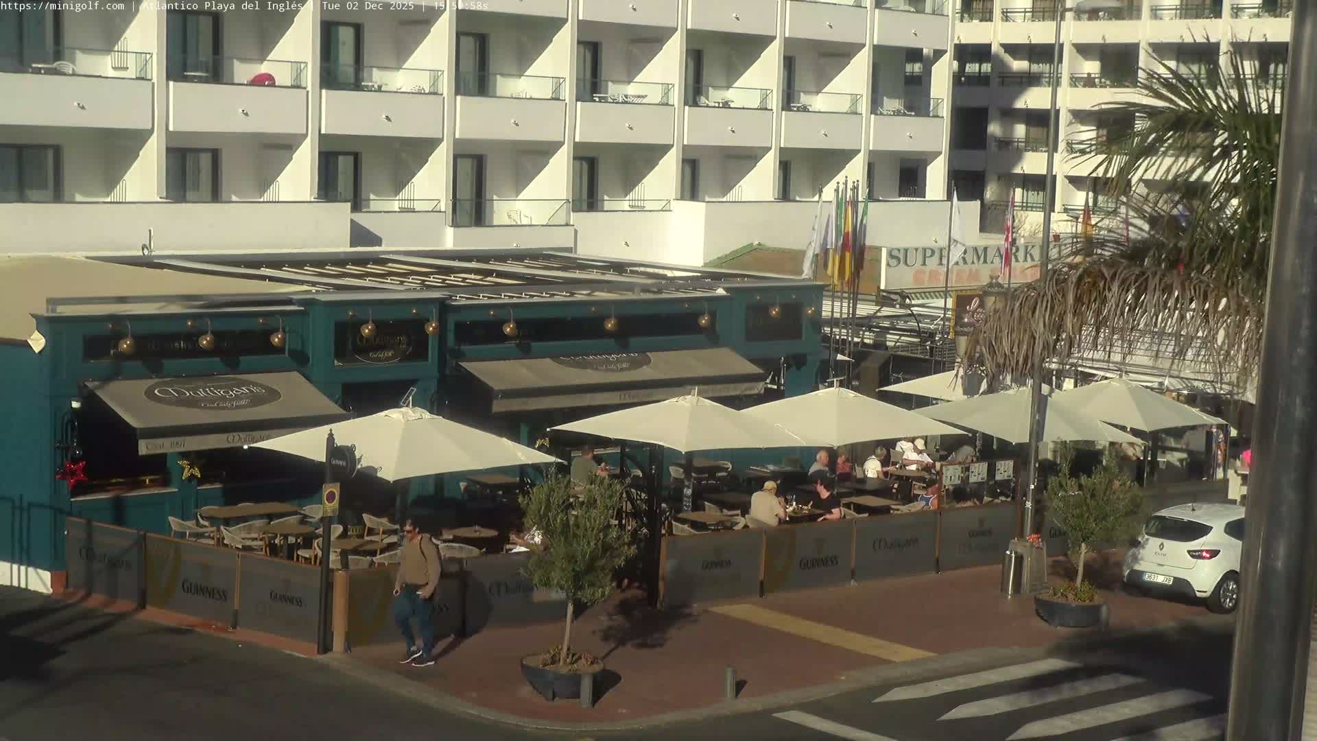 An outdoor pub or restaurant terrace is bustling with patrons seated at tables under large white umbrellas on a sunny day, set against a backdrop of a multistory white building and a prominent palm tree.
