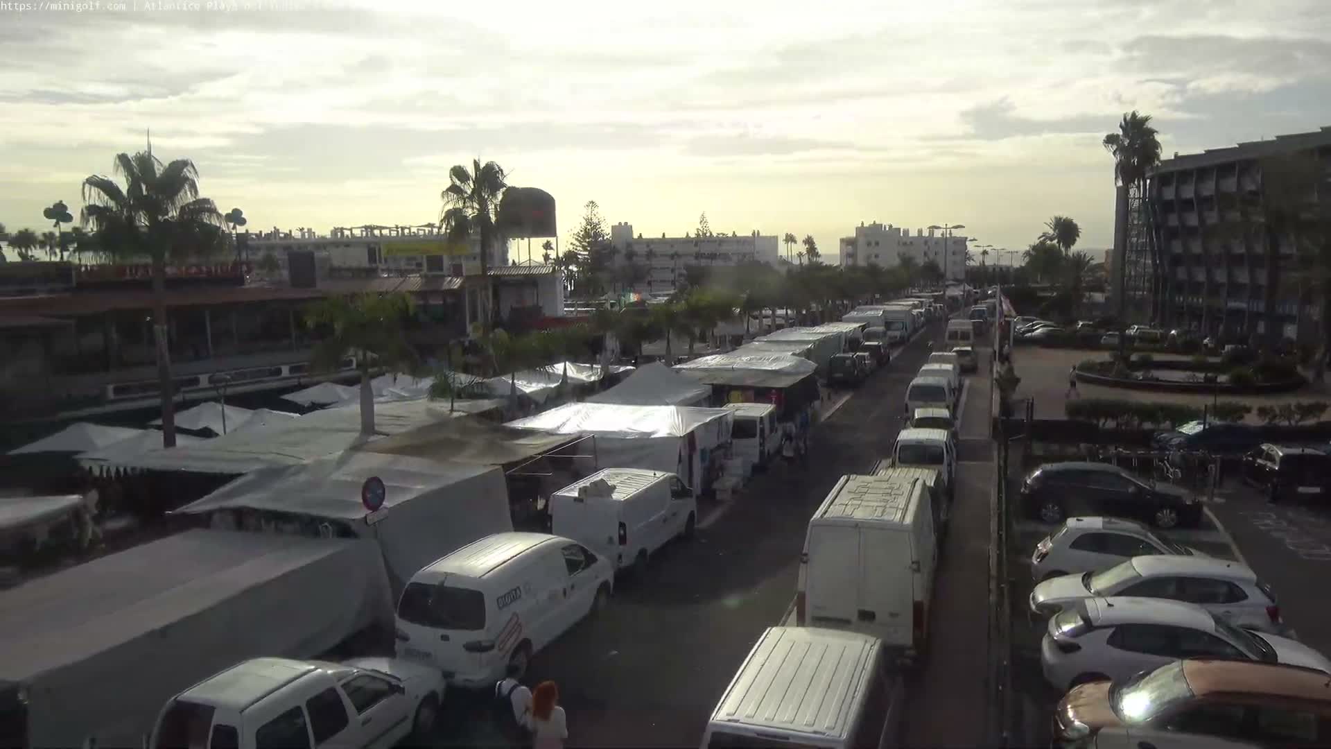 A bustling outdoor market street, lined with numerous white vendor tents, vans, cars, and palm trees, stretches towards distant buildings and the ocean under a bright, partly cloudy sky.