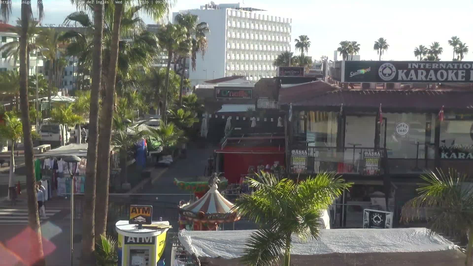 A bustling outdoor market street, lined with numerous white vendor tents, vans, cars, and palm trees, stretches towards distant buildings and the ocean under a bright, partly cloudy sky.