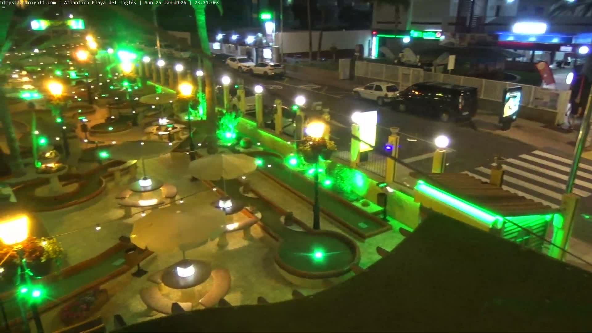 An outdoor mini-golf course is brightly illuminated at night with warm white and vibrant green lights, showcasing various holes, seating areas with umbrellas, and palm trees, set against a backdrop of a street with vehicles and commercial buildings under clear night conditions.