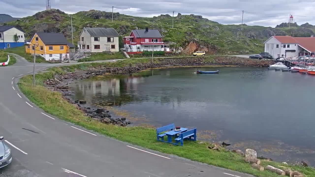 A calm, overcast day reveals a small harbor nestled between colorful houses and rocky hillsides, with a road curving alongside the water's edge.