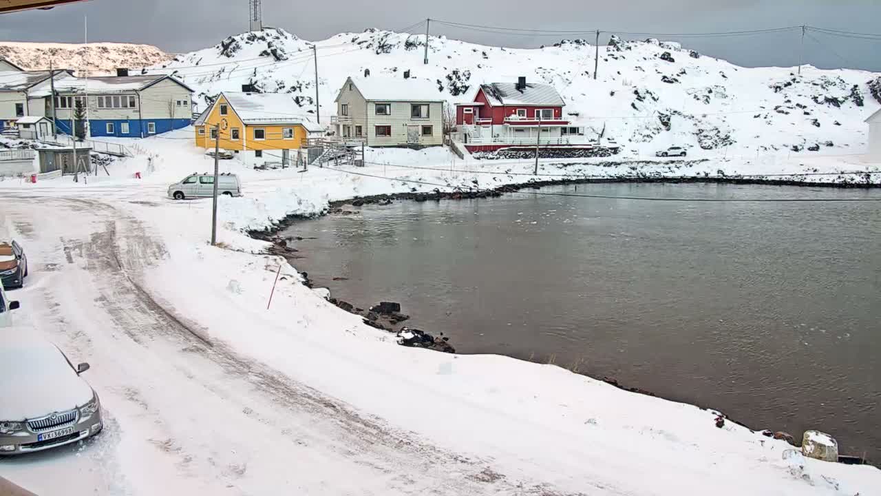 A snow-covered coastal village featuring colorful houses, a road with parked cars, and a dark body of water is seen under an overcast, wintry sky.