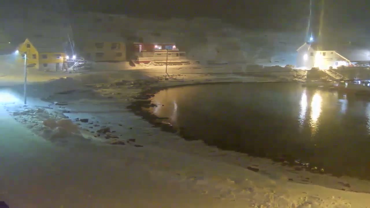 Several fishing boats are moored in a snowy harbor surrounded by snow-covered mountains and buildings under a cloudy, overcast sky, indicating cold winter conditions.