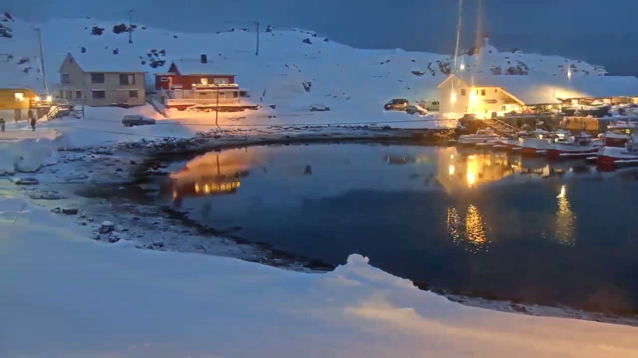 A snow-covered harbor is dotted with numerous fishing boats docked along the shore and framed by coastal buildings and distant snow-capped hills, all under an overcast sky with a glowing street light reflecting in the calm water.