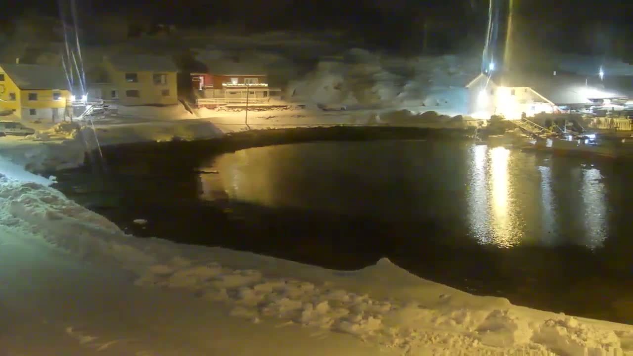A snow-covered landscape at night showcases brightly lit houses and structures along the edge of a dark body of water, which reflects their glow under a clear winter sky.
