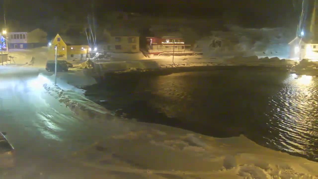 A snowy harbor scene features numerous fishing boats docked by brightly lit buildings with warm reflections on the water, all nestled among snow-covered mountains under an overcast sky.
