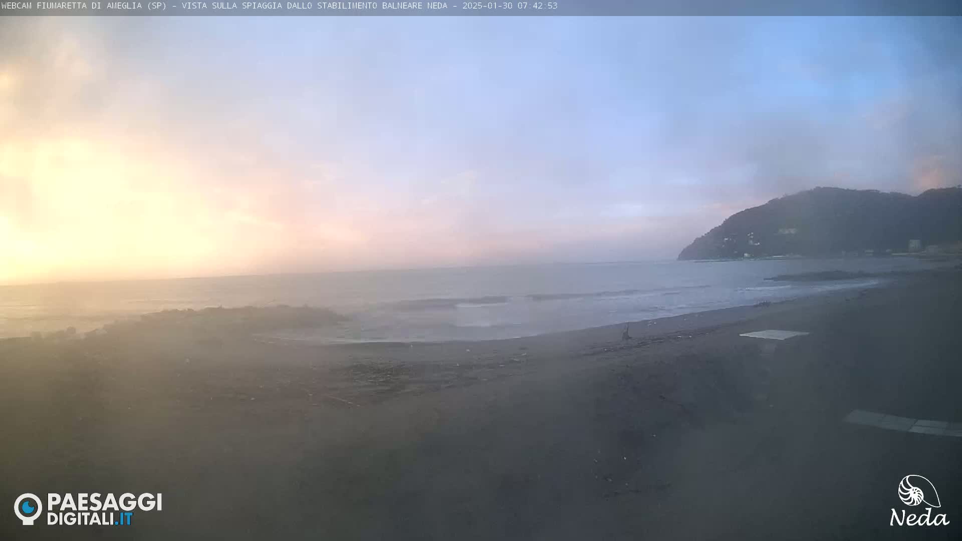 A hazy sunrise illuminates a dark sandy beach and a calm ocean, with a hill in the background.