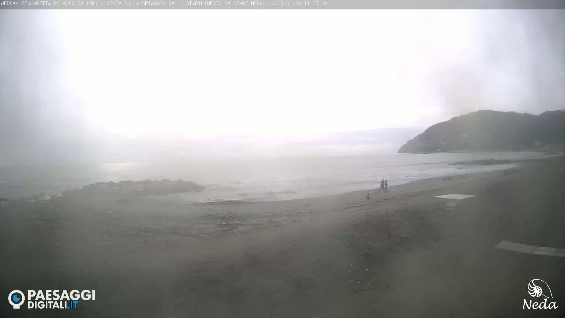 A foggy beach scene shows a couple walking along the shore, with a calm sea and a distant hillside visible in the hazy conditions.