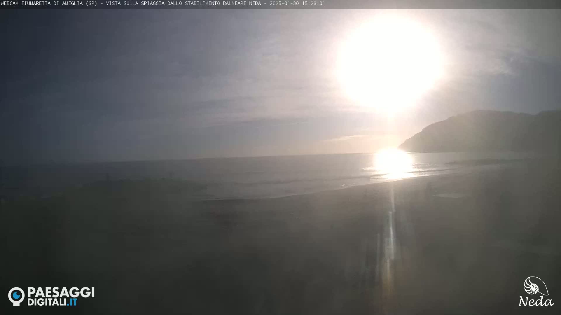 A hazy sun shines brightly over a calm ocean and sandy beach, with a dark hill visible in the distance.