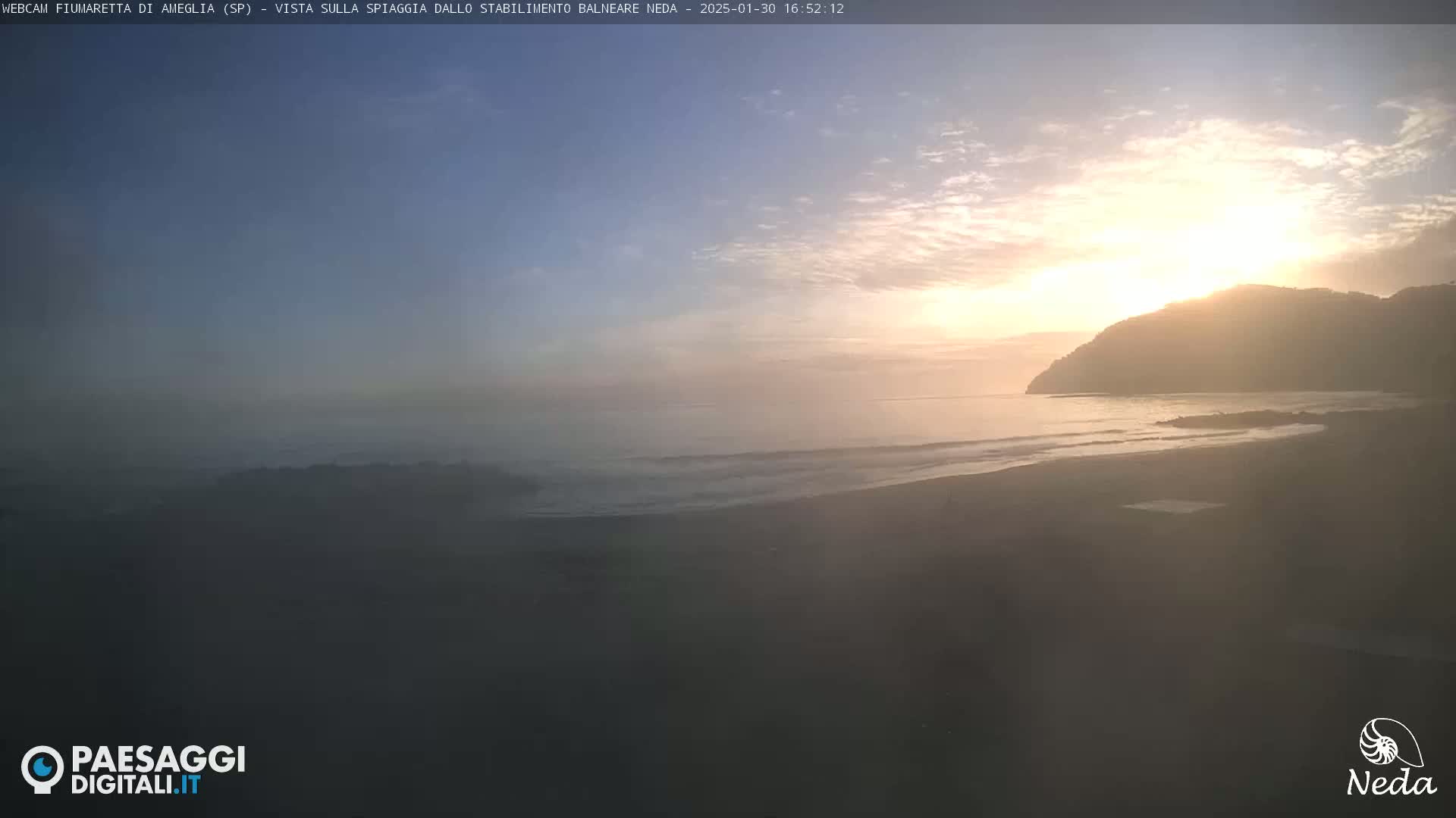A hazy sunrise illuminates a beach and ocean, with a hill visible in the background.