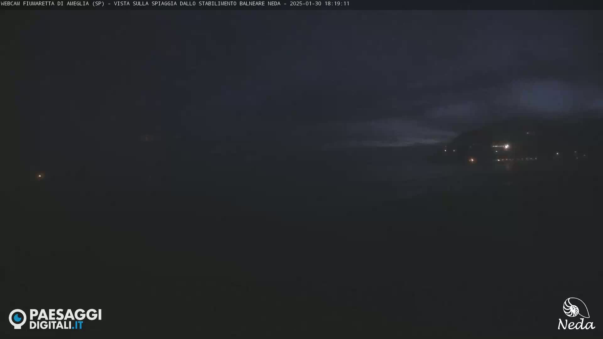 A dark, nighttime view of a beach with a dimly lit coastline under an overcast sky.