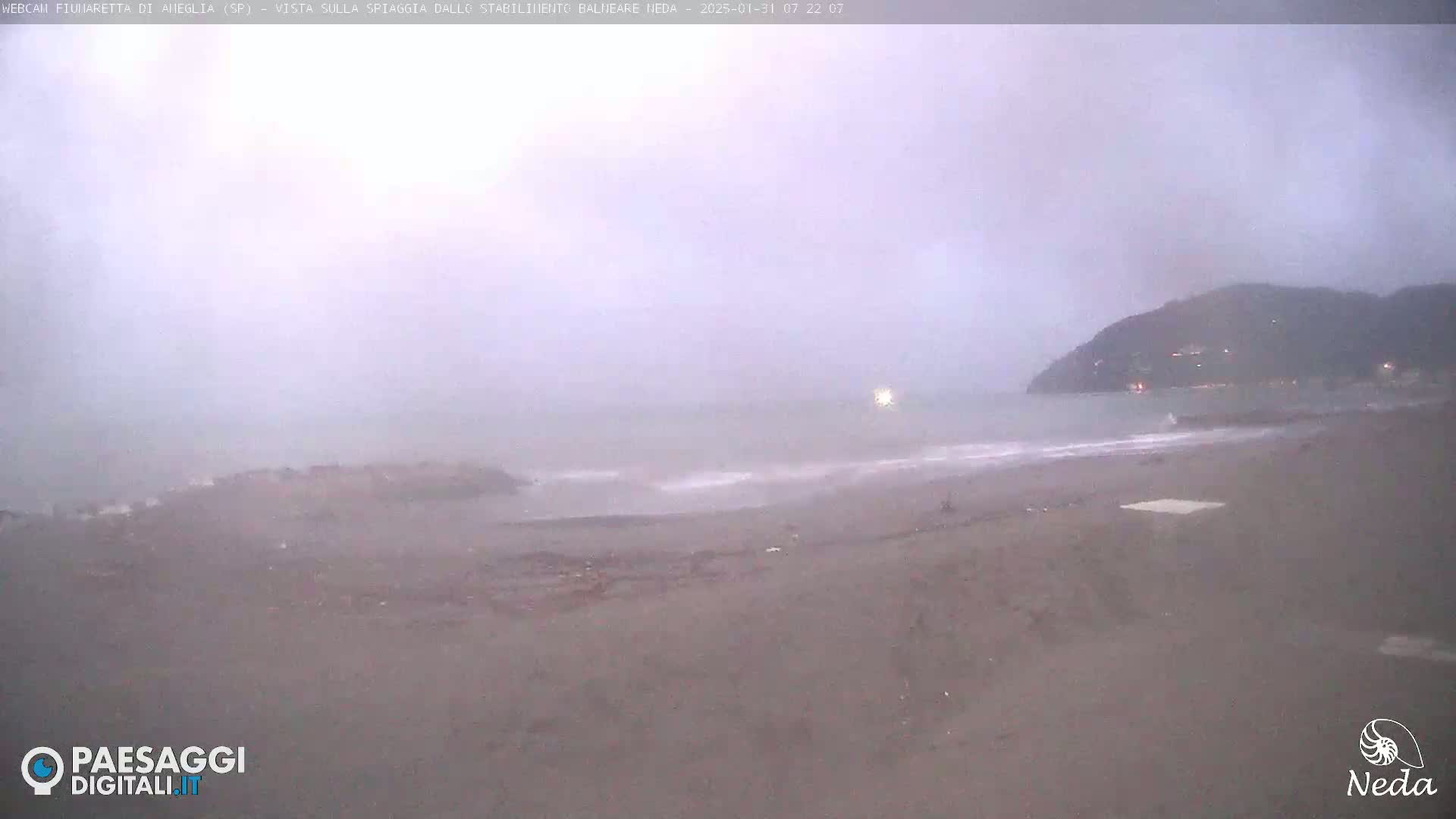 A hazy, overcast day shows a mostly empty beach with a calm sea and a distant hill in the background.