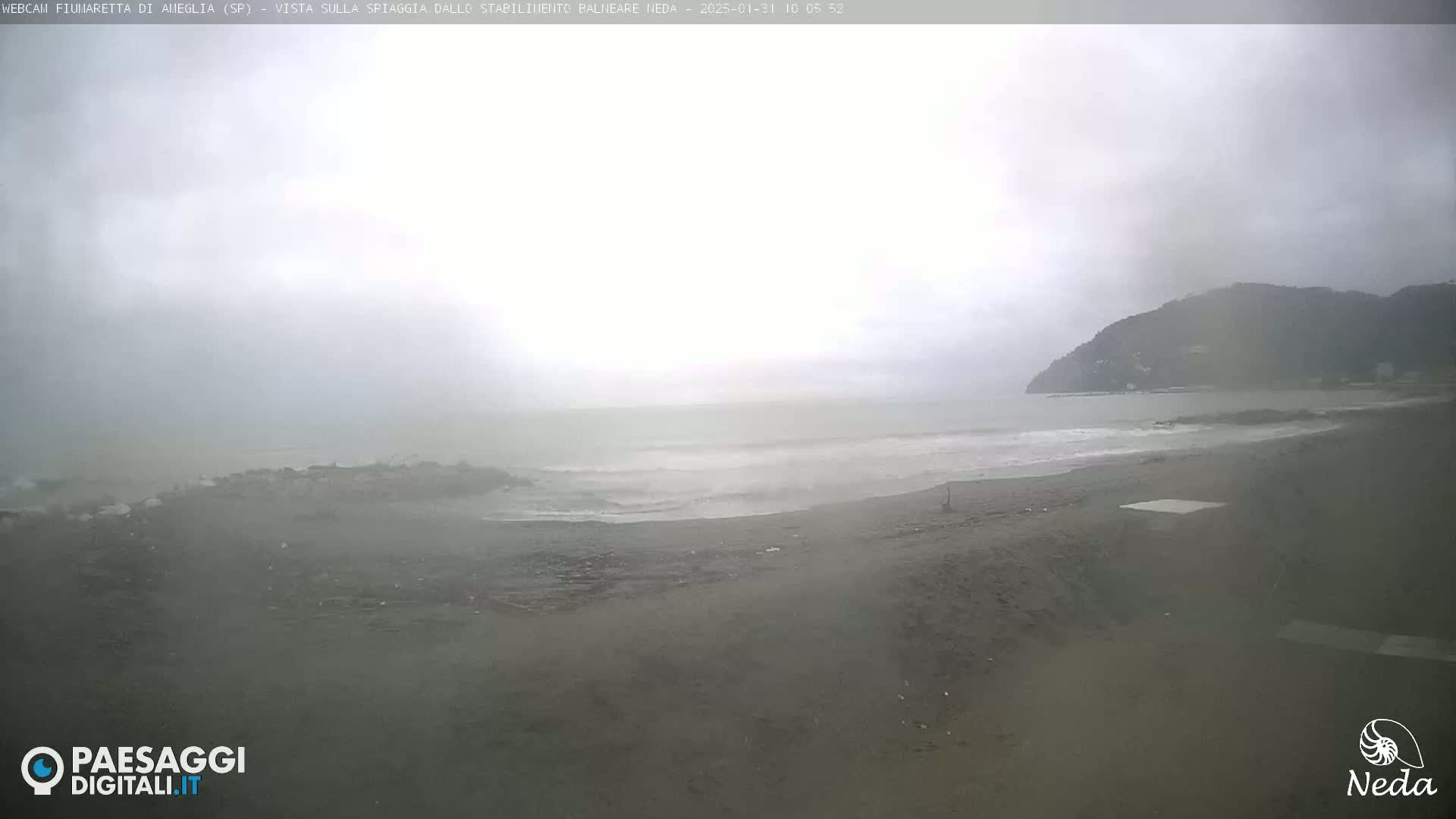 A mostly overcast, possibly rainy day shows a dark-grey beach with small waves lapping the shore, and a dark hill visible in the distance.