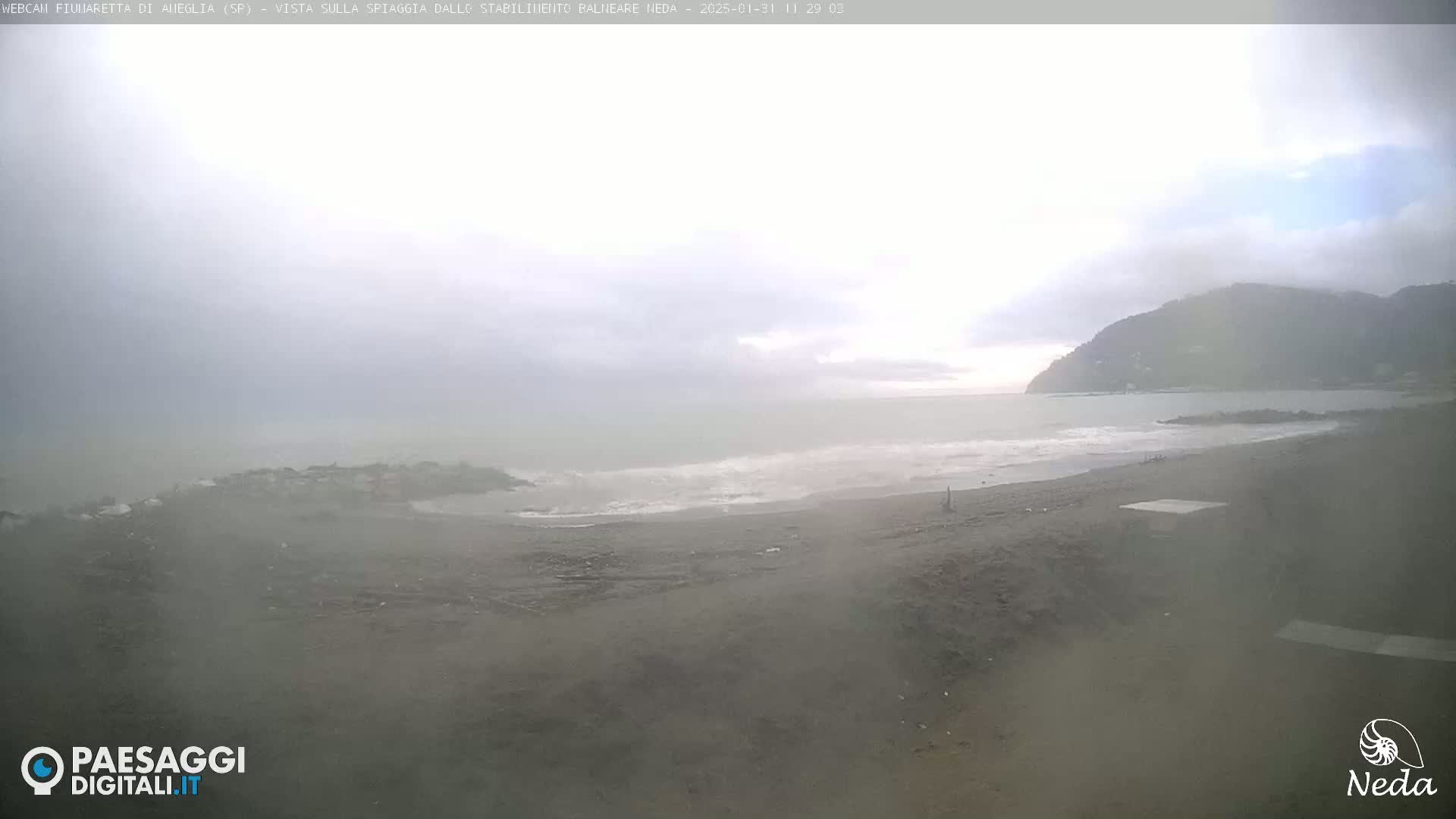 A hazy, overcast day shows a dark-sand beach meeting a gently wavy sea under a cloudy sky, with a distant, dark mountain visible in the background.