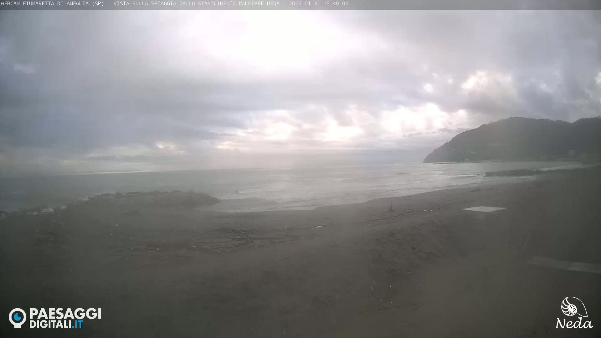 A dark, overcast day at a beach, with slightly wavy water and a distant mountain visible under a cloudy sky.