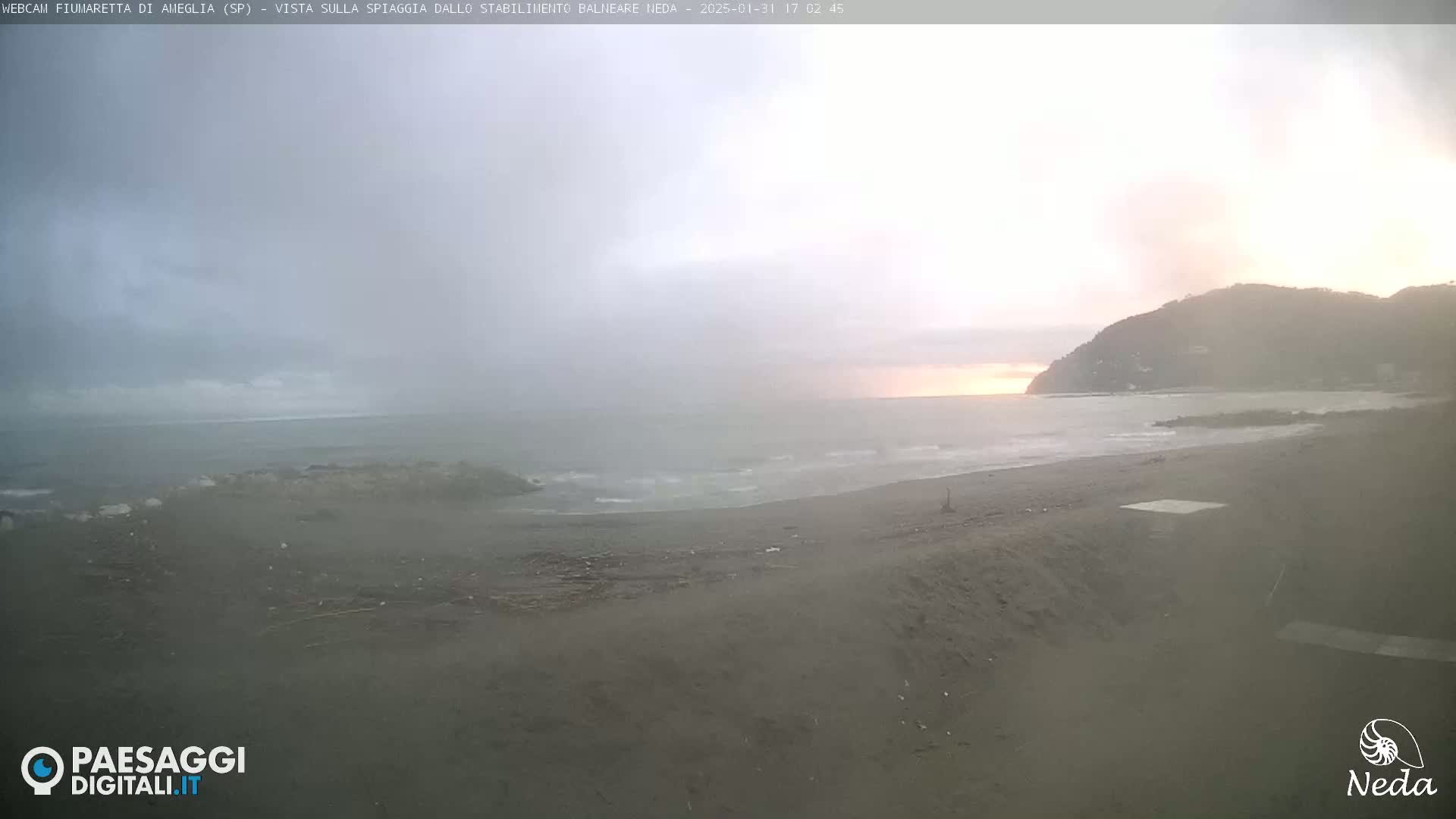 A hazy sunset over a dark-sand beach with gentle waves and a hill in the background.