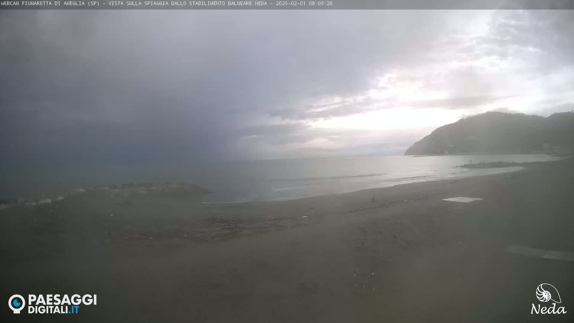 A hazy, overcast day shows a beach with a calm sea and a hill in the background.