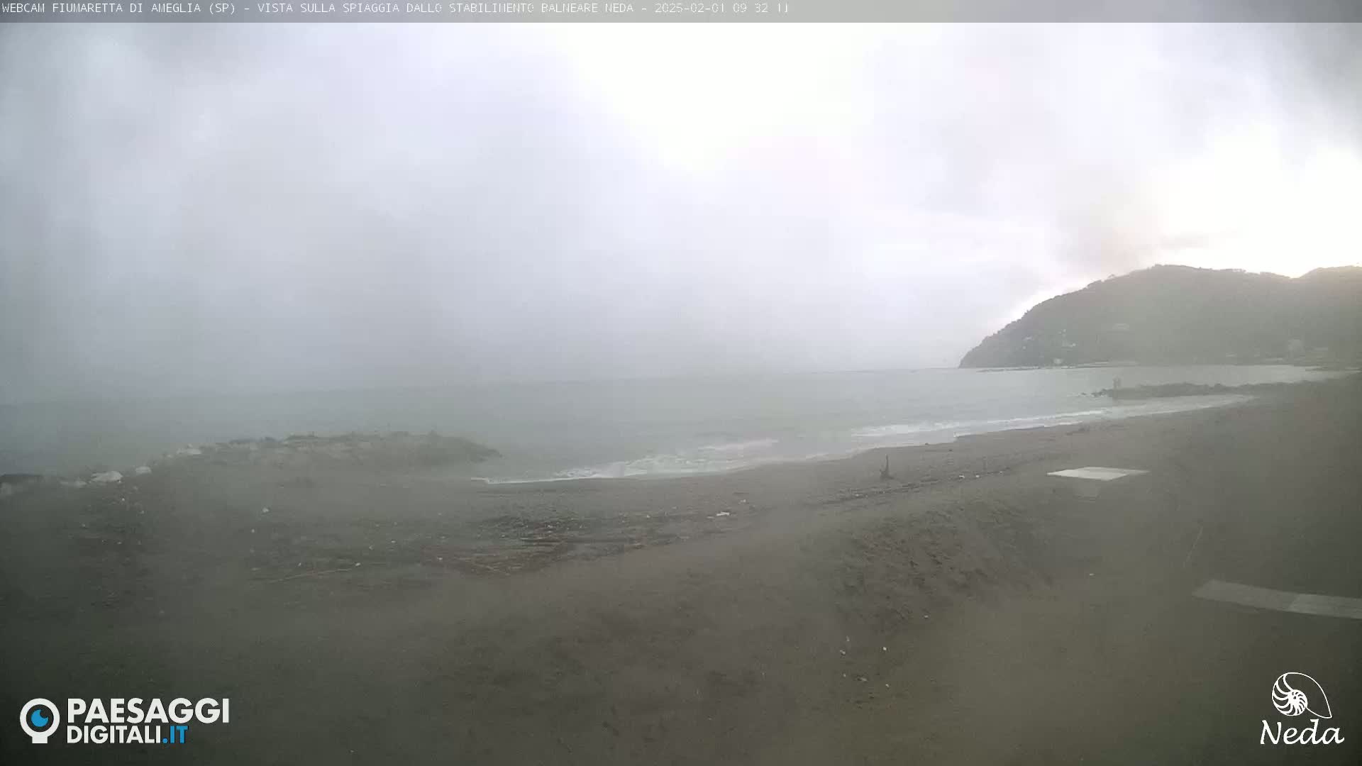 A foggy, overcast day at a beach, showing a grey sandy shore, a calm sea, and a hill in the distance.