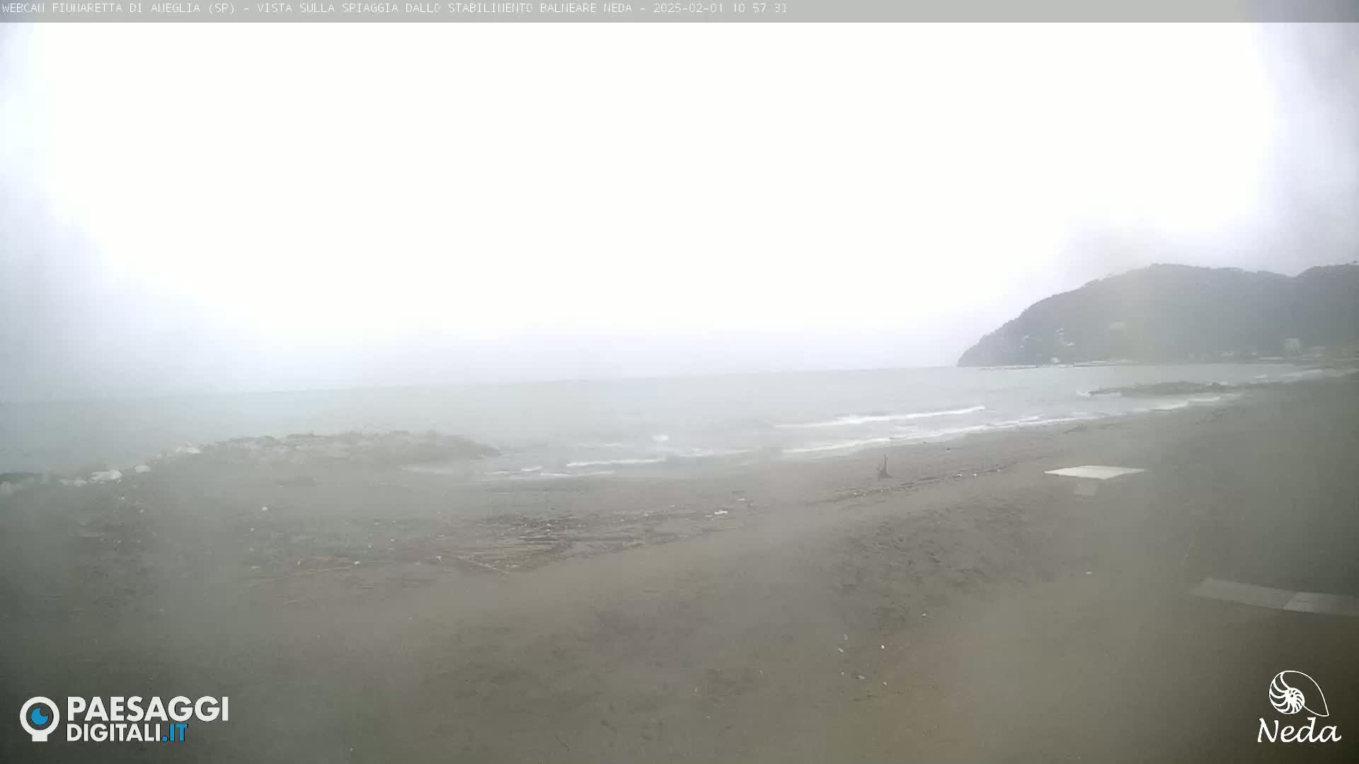 A hazy, overcast day at a beach shows a mostly empty, grey-sand beach with a distant, dark hill under a grey sky.