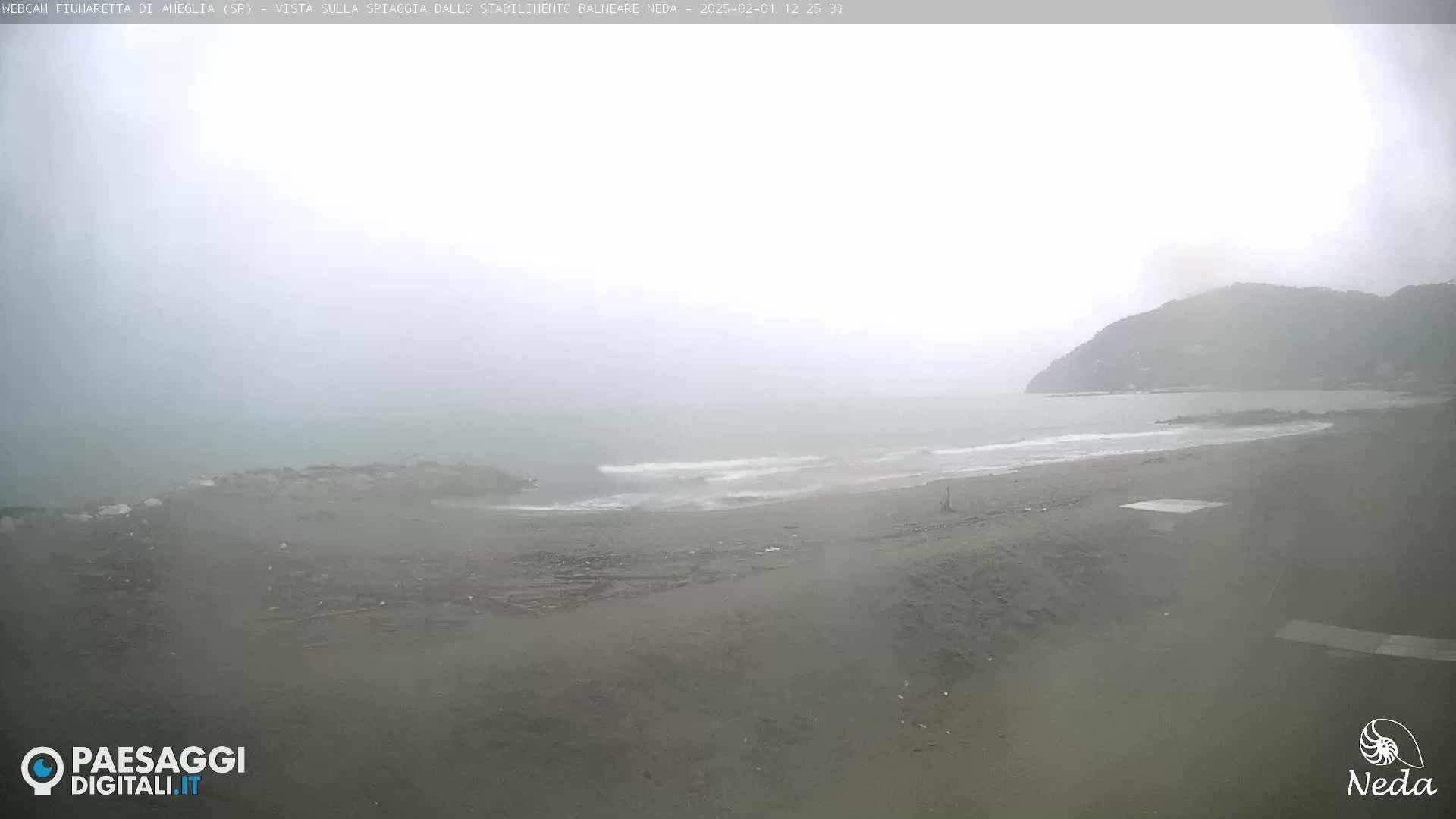 A hazy, overcast day shows a beach with small waves lapping the shore and a hill in the background.