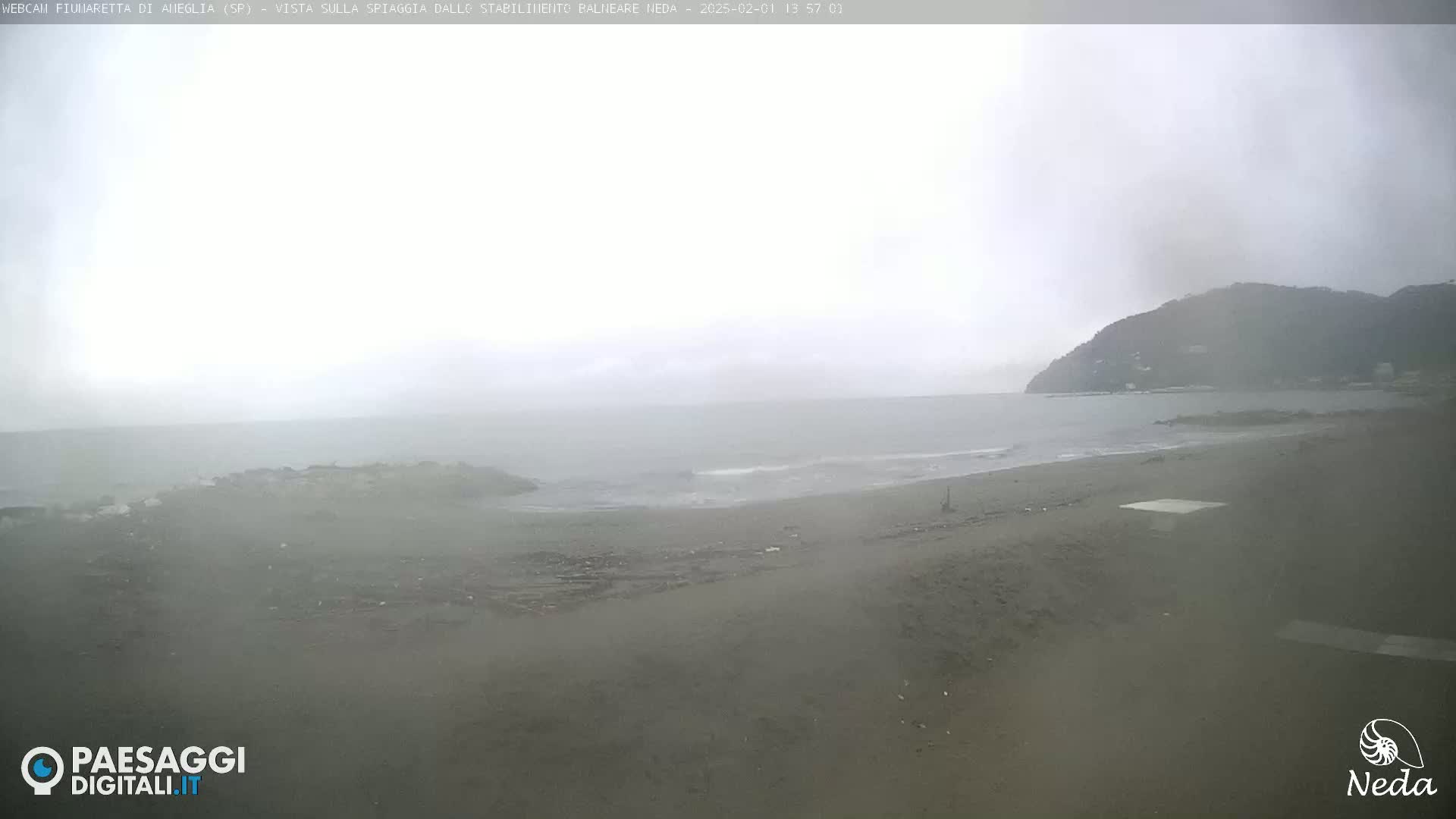 A beach with a calm sea and a hazy, overcast sky is visible from a high vantage point, showing a dark, sandy beach and a distant hill.