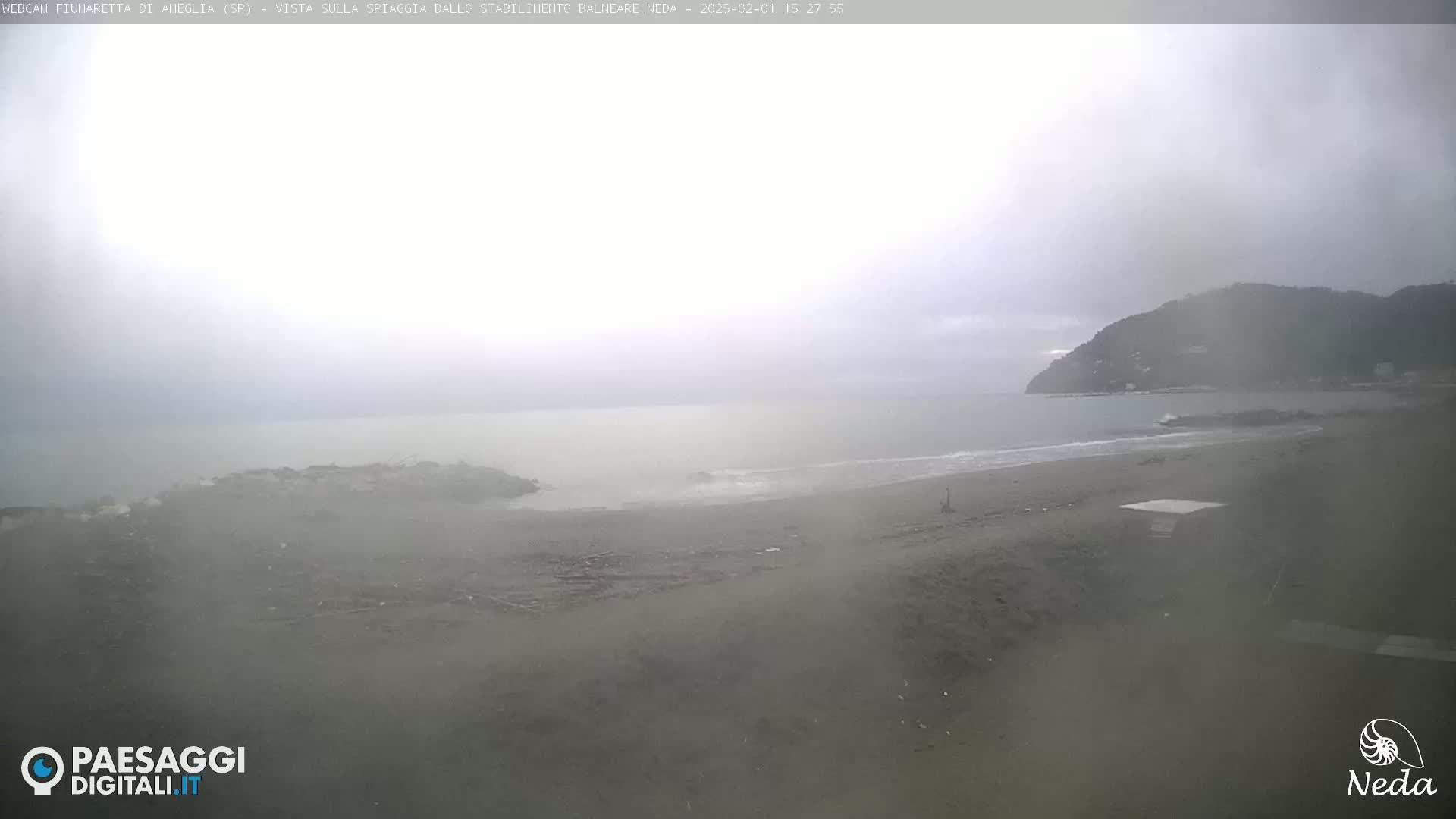 A hazy, overcast day shows a beach with a small structure and distant hills across a calm sea.