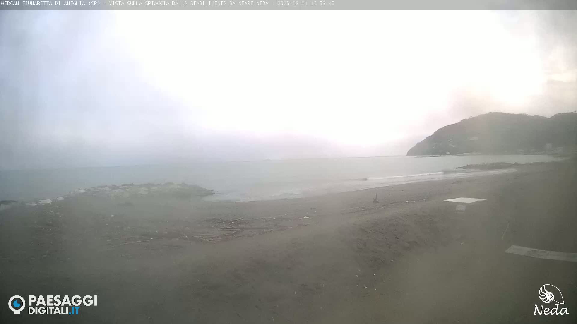 A hazy, overcast day shows a sandy beach and calm ocean with a distant hill in the background.