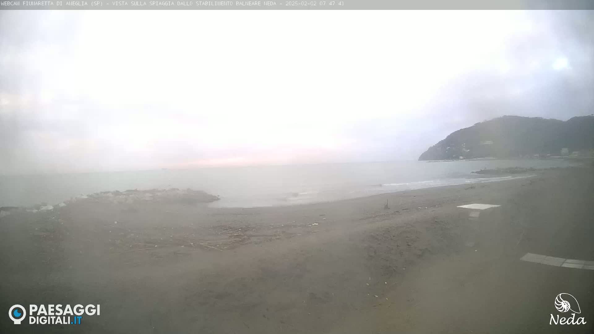 A hazy, overcast day at a sandy beach with a calm sea and a distant mountain range.