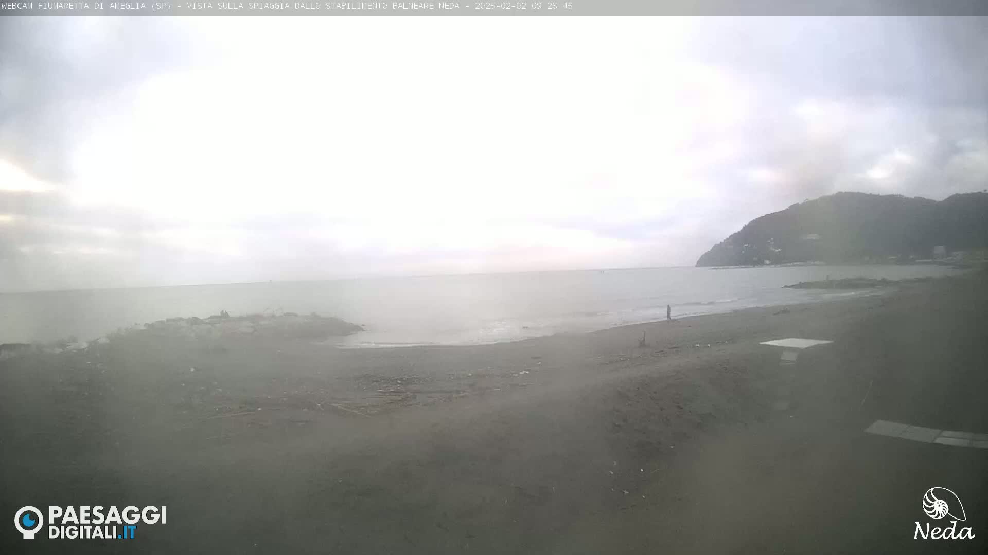 A hazy, overcast day at a beach with a dark sand shoreline, calm sea, and a hill in the distance.