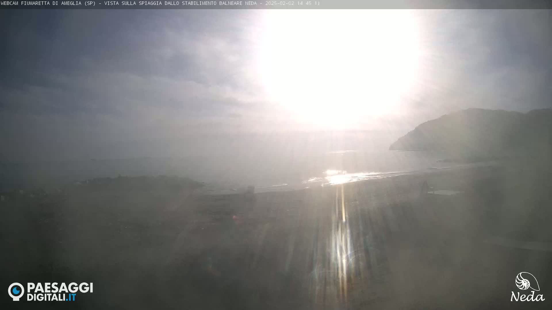 A hazy, bright sunny day overlooks a beach and calm ocean with a hill in the distance.