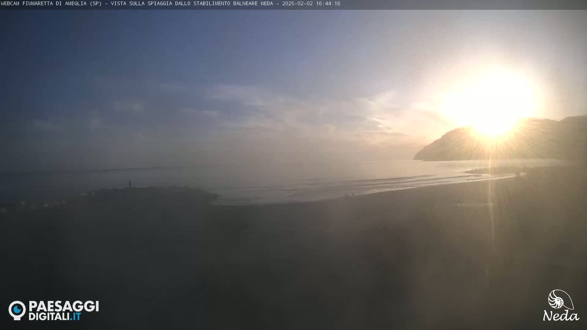 A hazy sunrise illuminates a dark sandy beach and calm ocean, with a distant hill silhouetted against the bright sun.