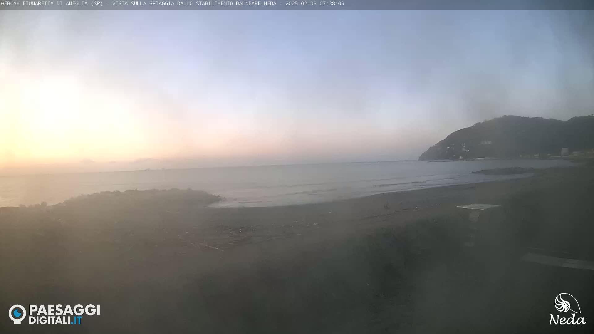 A hazy sunrise over a calm sea and sandy beach, with a dark hill visible in the distance.
