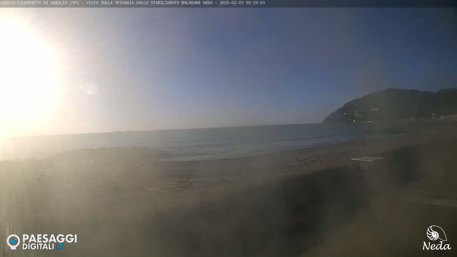 A hazy sunrise illuminates a beach and calm ocean with a distant hill in the background.