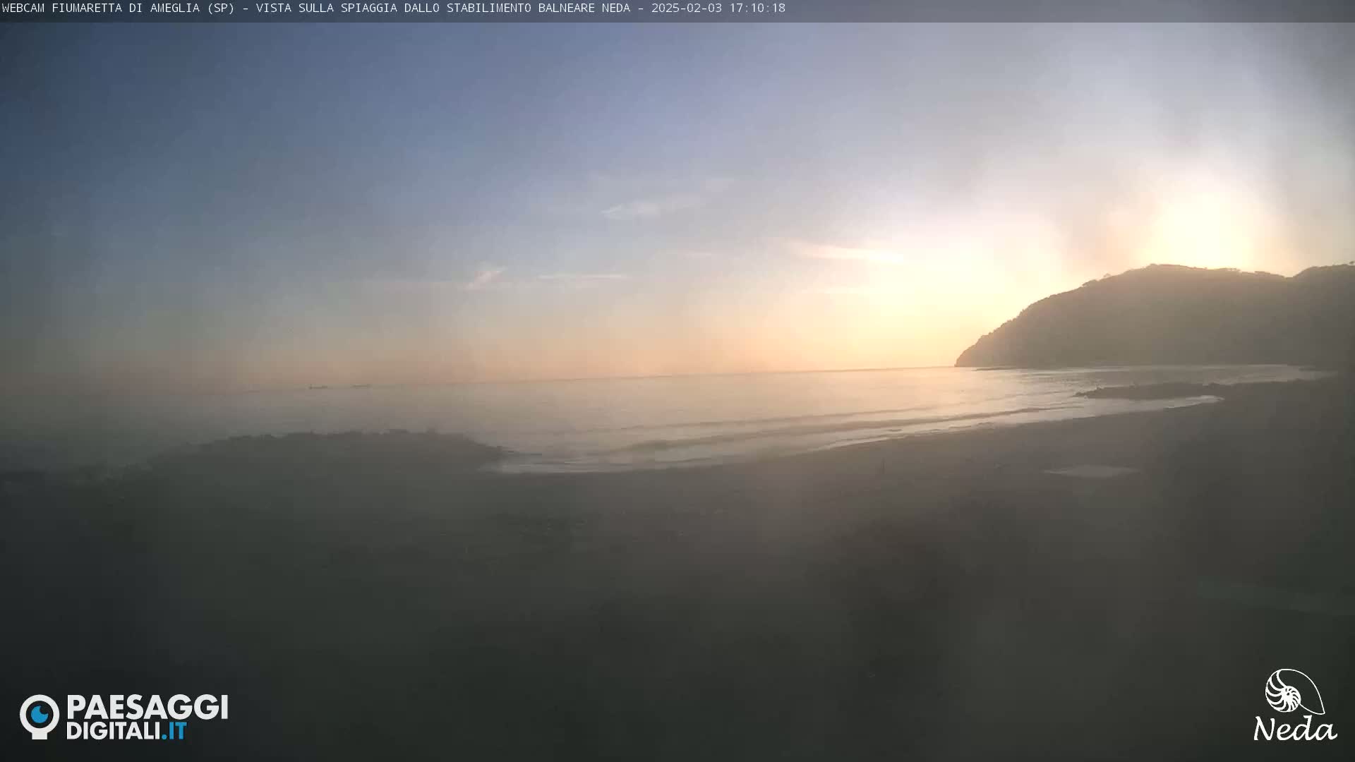 A hazy sunrise casts a warm glow over a calm ocean beach backed by a dark hill.