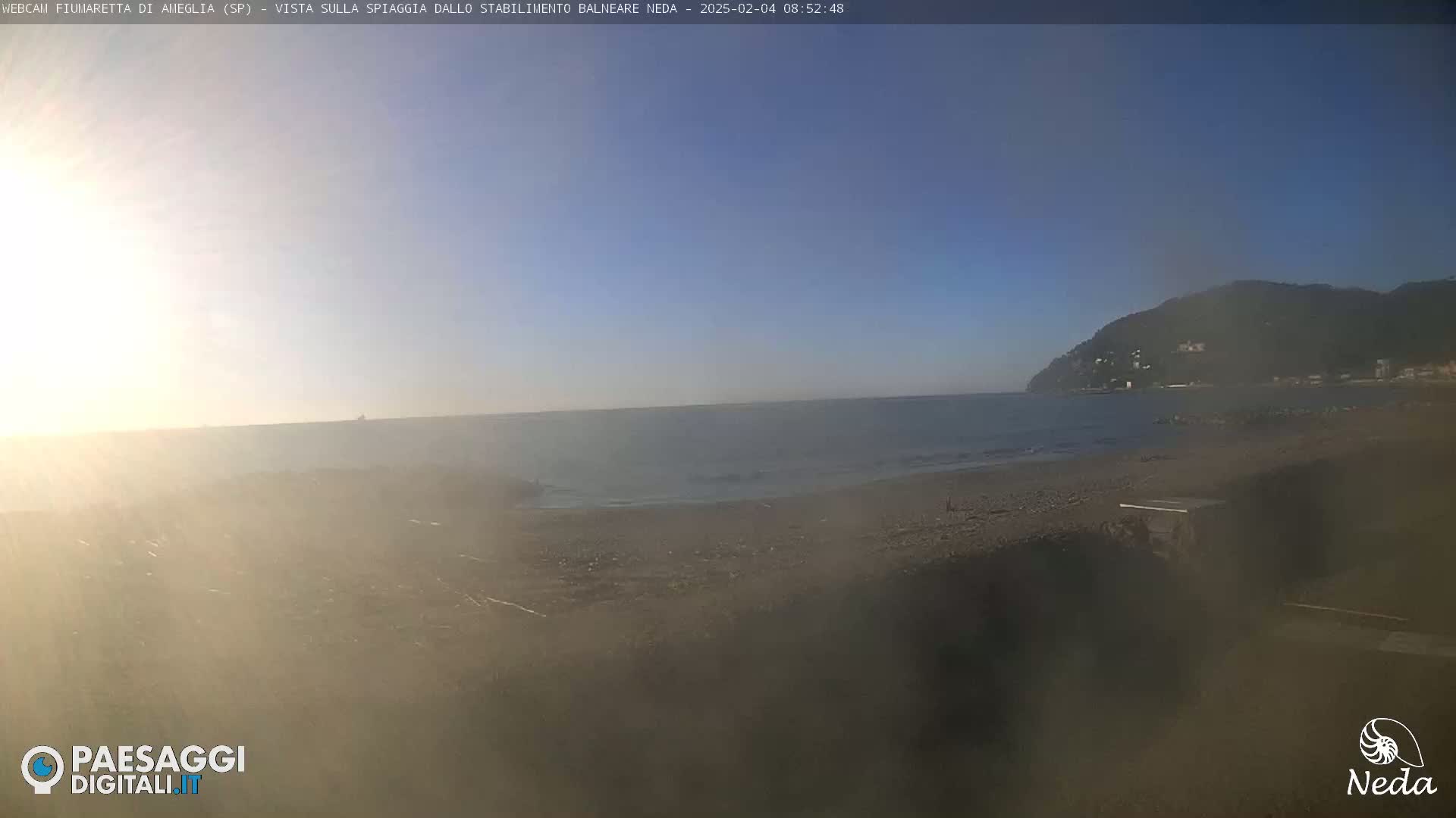 A hazy sunrise over a calm sea and sandy beach, with a distant mountain range visible under a clear blue sky.