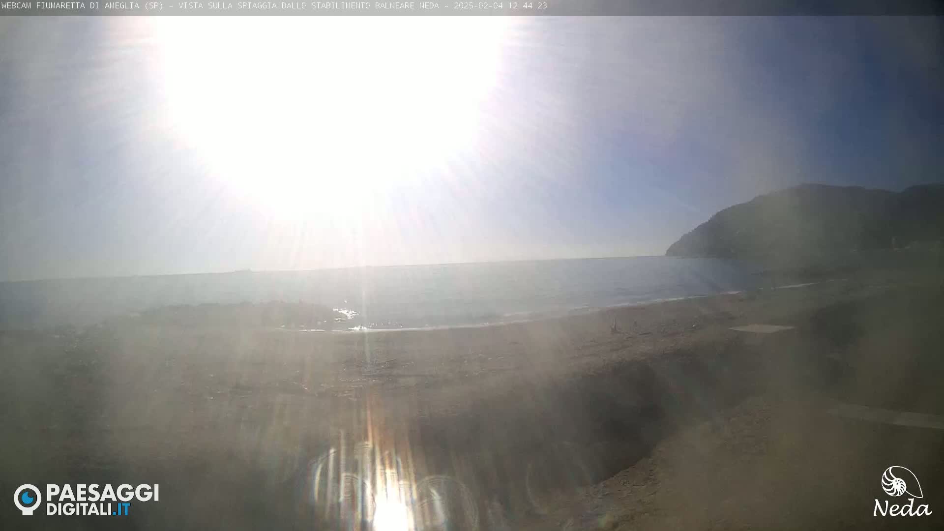 A sunny beach scene shows a calm ocean meeting a sandy shore, backed by a distant hill.