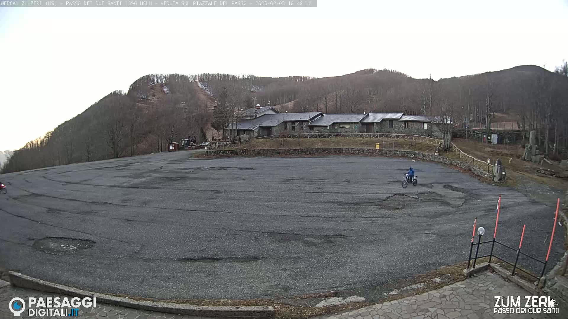 A large, empty asphalt parking lot is situated in front of a cluster of stone buildings, with a person on a dirt bike in the center, all set against a backdrop of a snow-dusted hillside under a mostly clear sky.