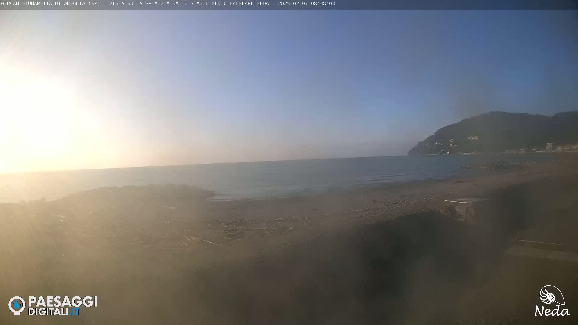 A hazy, sunny day at a beach with calm water, showing a shoreline with dark sand and a hill in the distance.