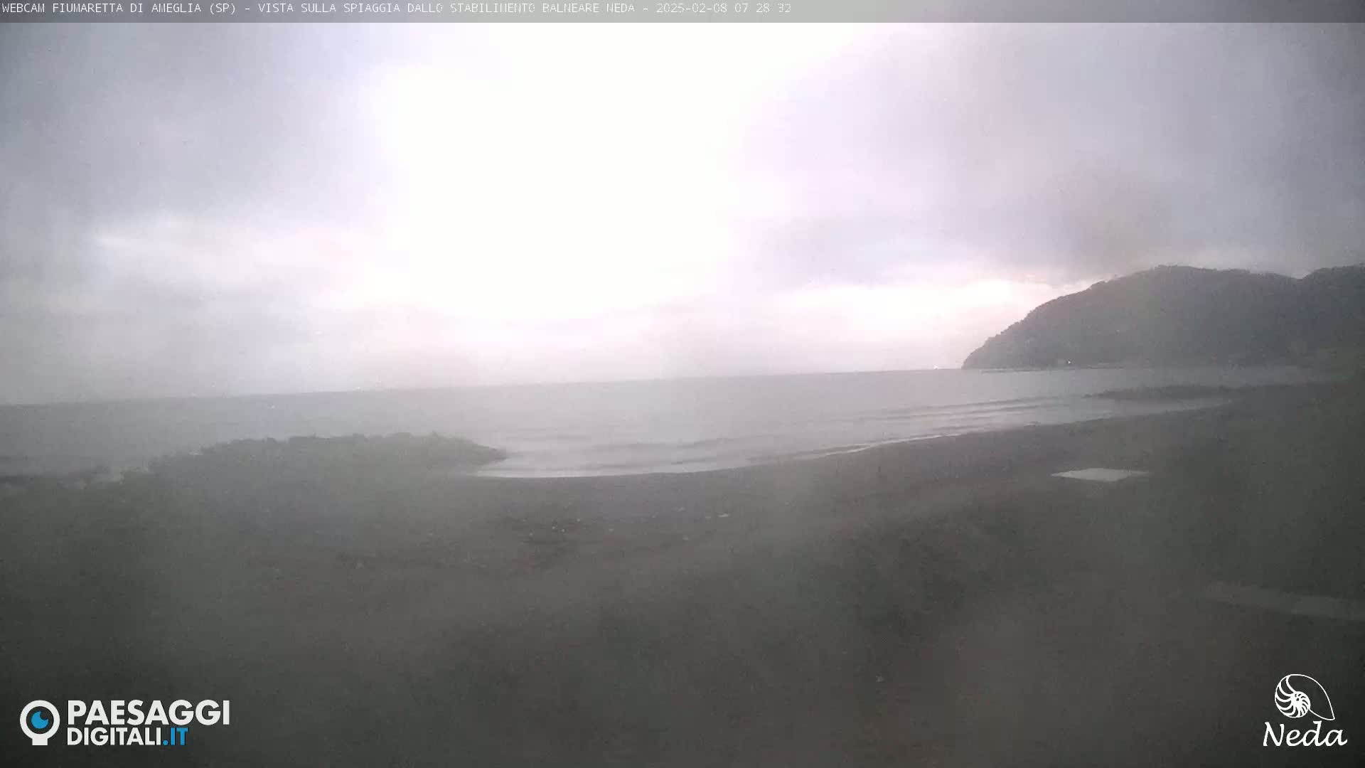 A dark, overcast day shows a view of a dark-sand beach and calm ocean waves lapping the shore, with a distant hill in the background.
