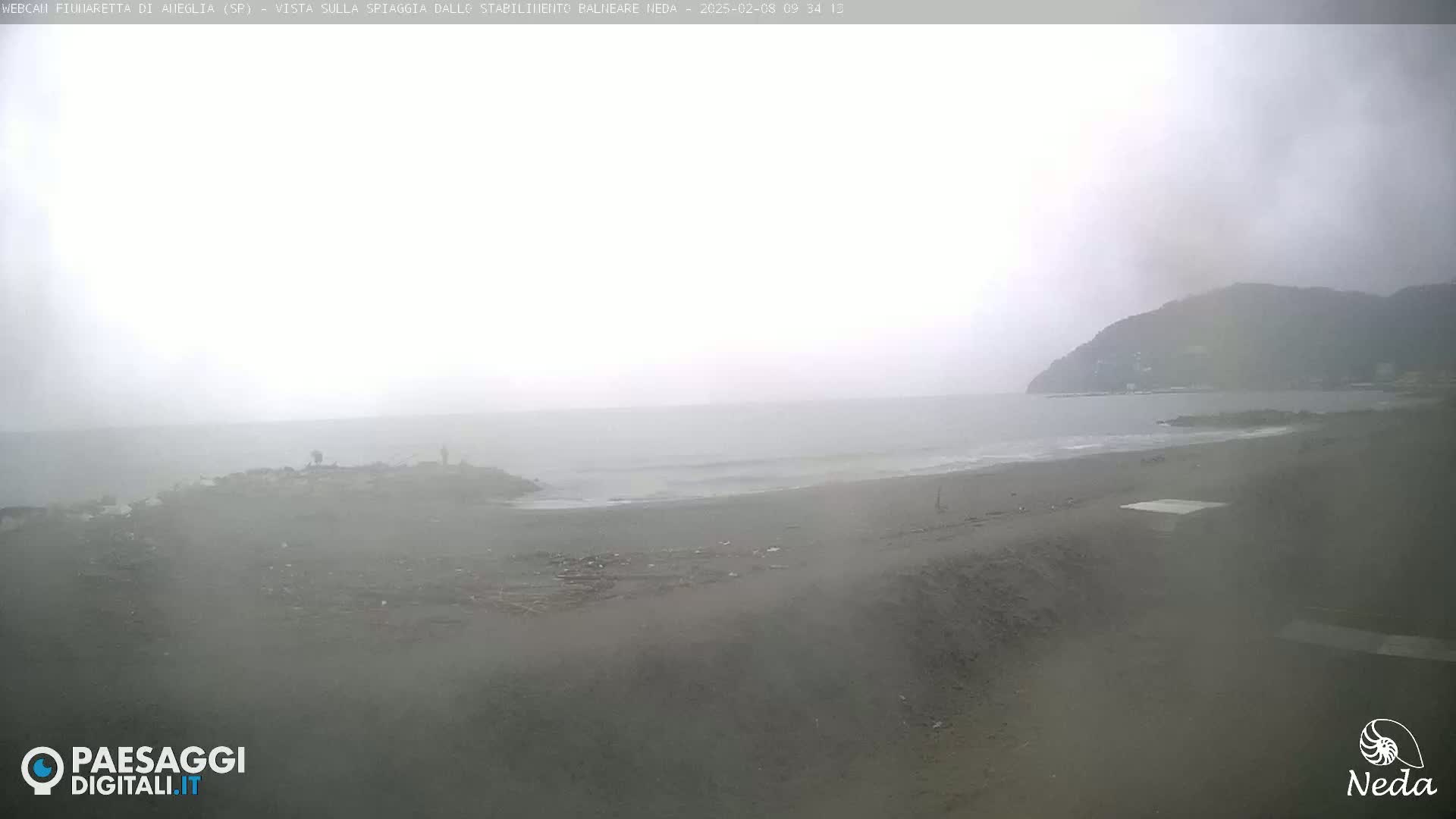 A hazy, overcast day reveals a dark-sand beach meeting a calm, gray sea, with a distant hill visible in the background.