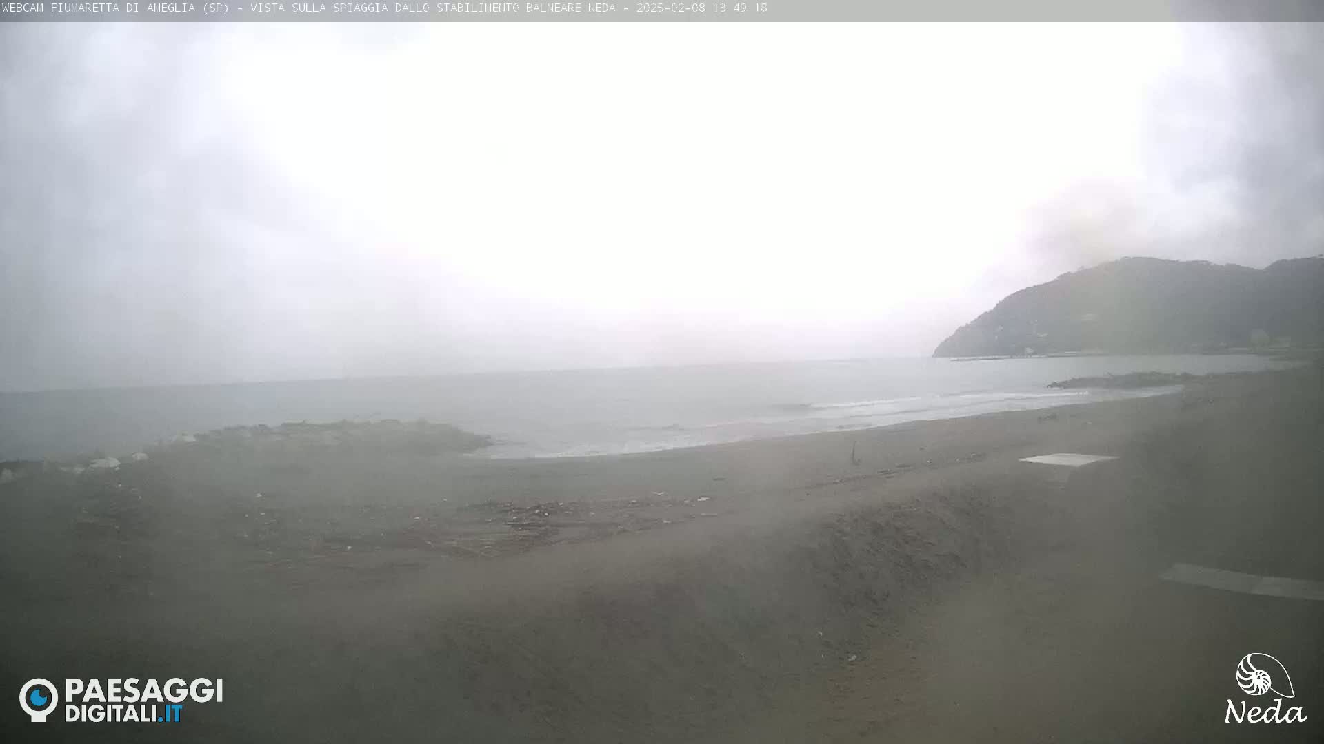 A hazy, overcast day reveals a beach and calm ocean with a distant hill visible through the mist.