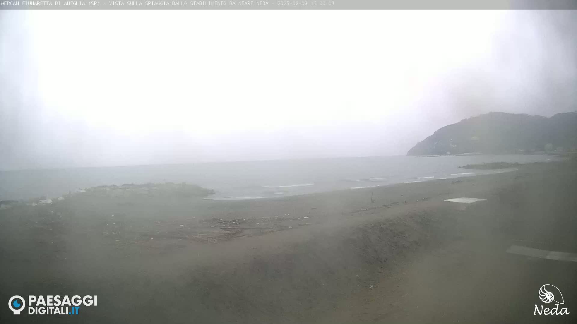A hazy, overcast day shows a beach scene with a calm sea and a hill in the background.