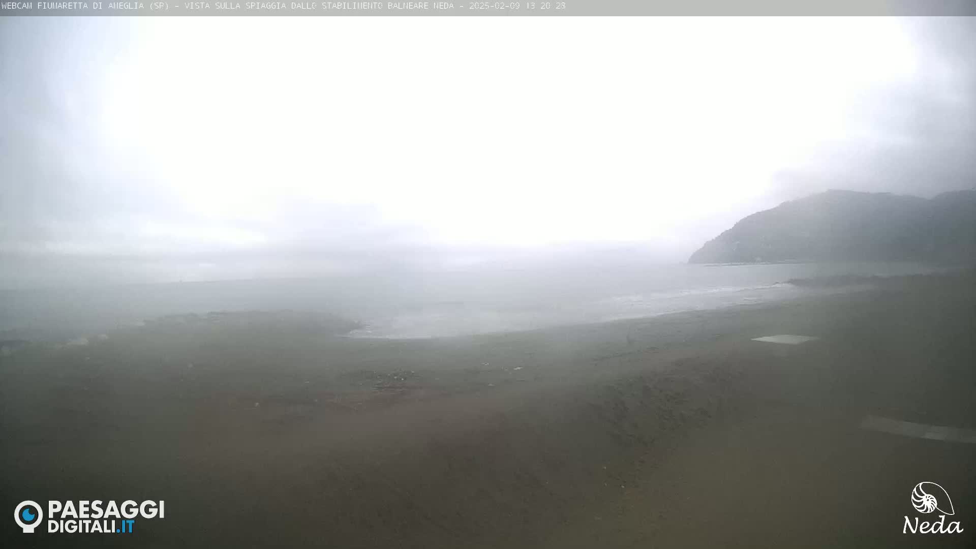 A hazy, overcast beach scene shows a mostly gray shoreline and a distant, indistinct mountain under a foggy sky.
