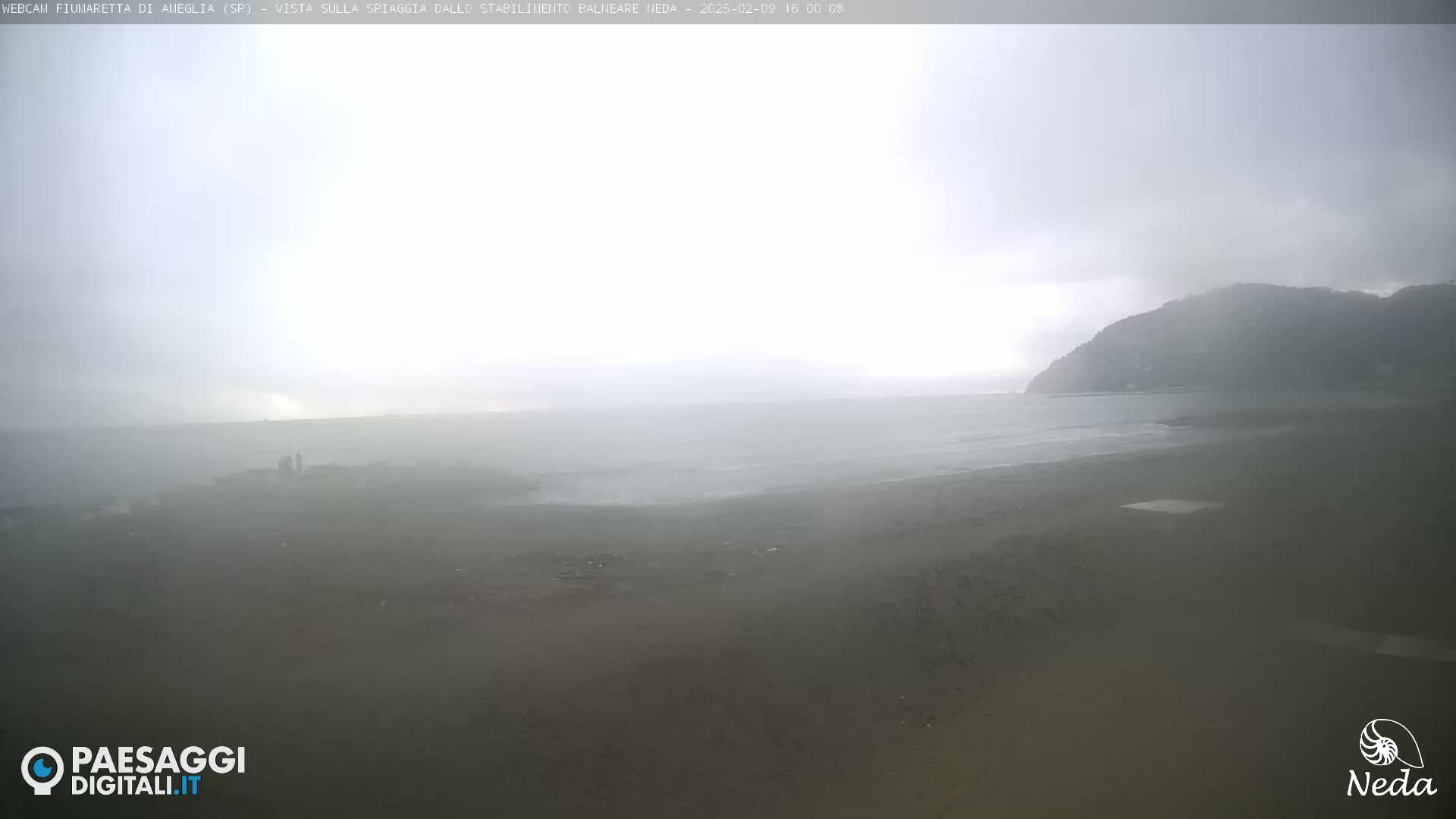 A hazy, overcast day reveals a beach scene with a calm sea and a distant hill shrouded in mist.