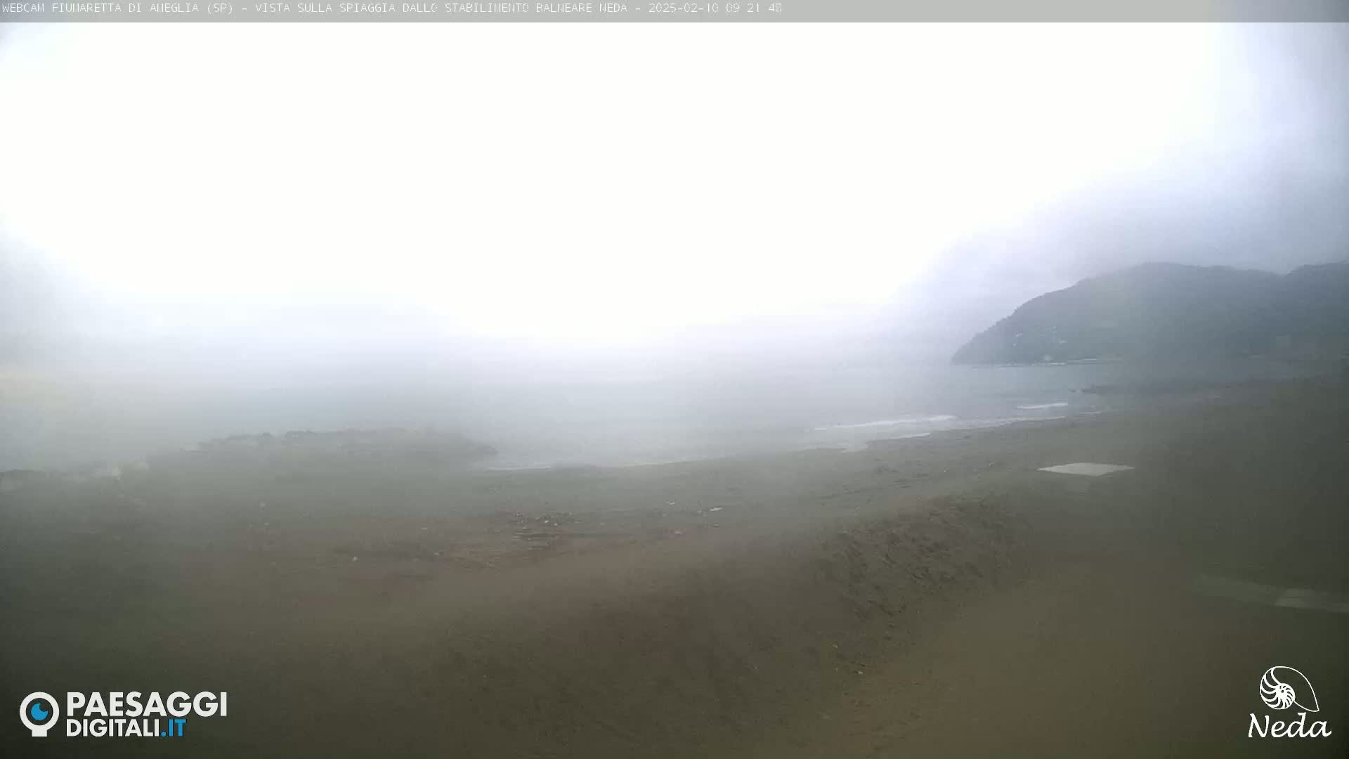 A foggy beach scene shows a sandy shoreline and calm water extending to a distant, hazy mountain range.